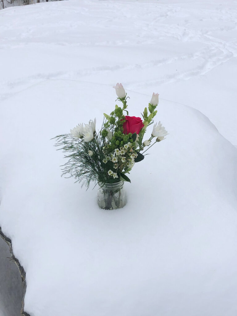On a snow-covered granite table, a vase filled with evergreens, white flowers, and a single red rose