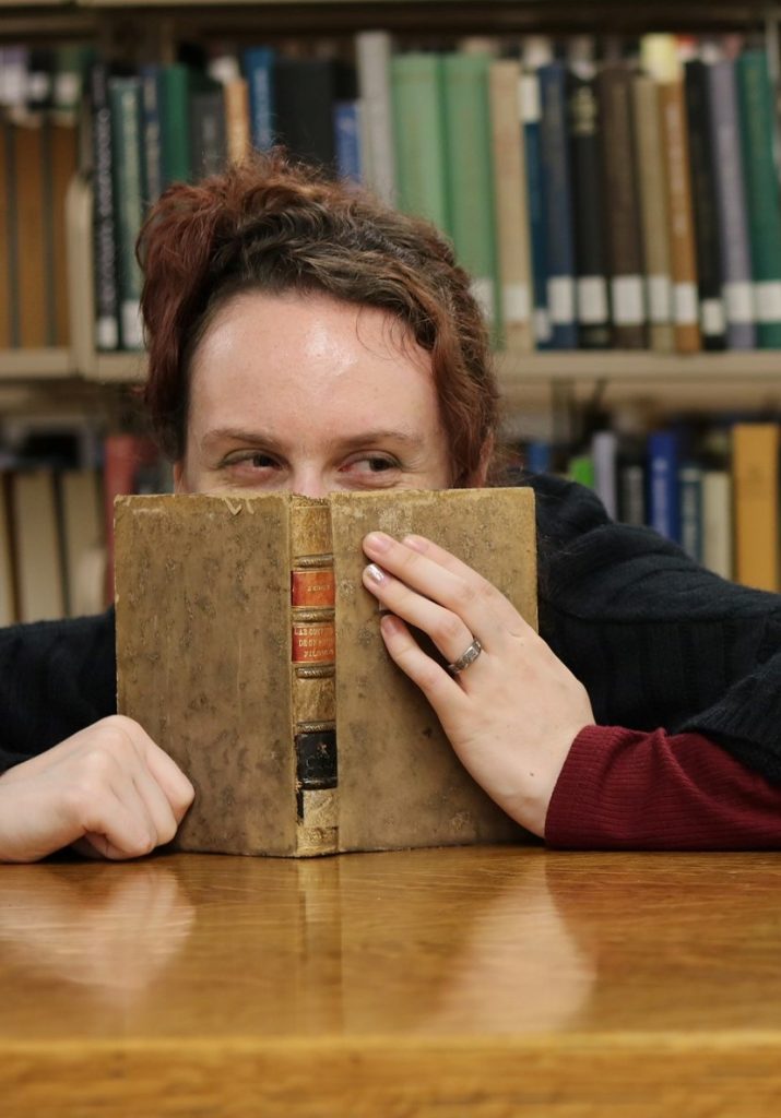 A brilliant, kind, and mischievous woman with more than a hint of Lyaquendi ancestry peeks at us over the edges of an ooooold book. She's in a library (duh) and her short reddish spiky hair matches both the sleeves of her shirt and some of the books behind her which is just fabulous.